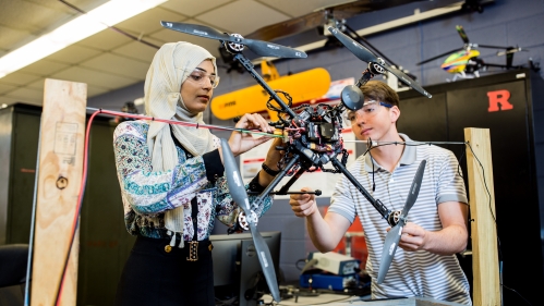 A male and female student work on a drone in a lab. The female is on the left, wearing black pants and a multi-colored top and a headscarf, the male to the right has brown hair and is wearing a gray polo shirt.