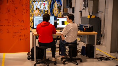 A female and male student sit at a desk with their backs to the camera. They are in a lab looking a desktop computer screens. To the left is an orange board with mathematical equations.