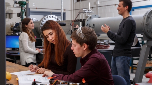 Four college students work in a lab. In the foreground and male student and female student consult a text book at a lab bench. In the background a male student and female student pose near a wind tunnel device.