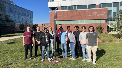 11 engineering students stand holding a rocket outside