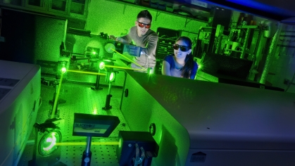 Male and female students working in a laser lab. They are wearing protective glasses and the room is tinted green from the lasers.