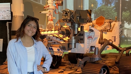 Young college student stands with one hand on her hip posing in front of a lunar rover. She is wearing jeans, a light blue blouse unbuttoned over a white shirt. She has long dark hair.