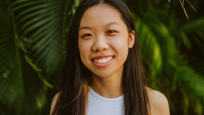 Female college student poses outside in front pine trees. She has long dark hair and is wearing a white sleeveless top.