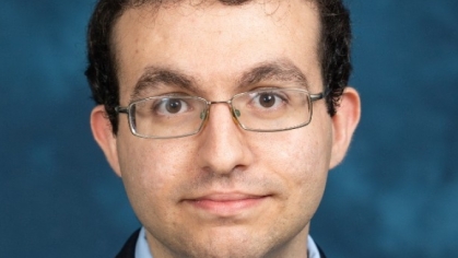 Head shot of male with dark brown, short hair wearing eyeglasses, a dark suit with a light blue button down shirt with a patterned tie. and