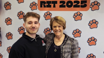Male student standing on left side and female professor standing on right side in front of a photo backdrop with orange paw prints