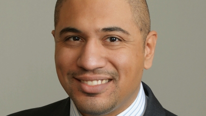 headshot of man with white and blue stripe collared shirt, blue, black, and grey, tie with a black jacket