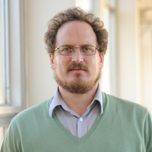 Head shot of male with brown hair, beard, a moutache, and eyeglasses, wearing a green v neck pullover sweater and a collared button down shirt.