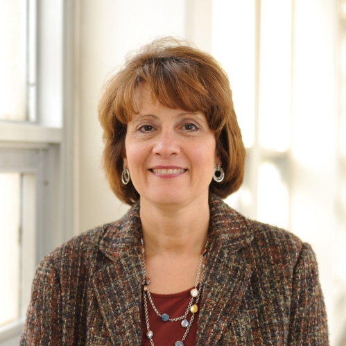 Head shot of woamn with brown hair, wearing a brown suit jacket, a maroon dress, necklace and earrings.
