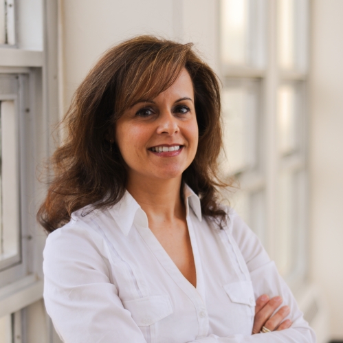 Head shot of female with her arms folded with long brown hair, wearing a white blouse
