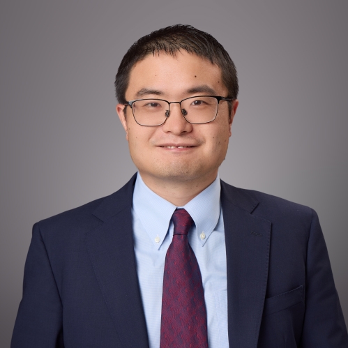 Headshot of male with short black hair and eyeglasses wearing a dark blue suit jacket, a light blue button down shirt, and a burgundy tie.