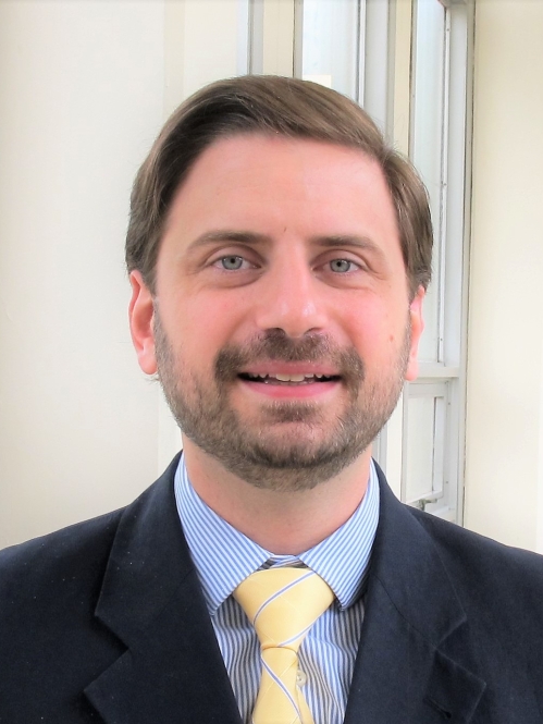 Head shot of male with brown hair, moustache, and beard, wearing a dark suit jacket, with blue button down shirt, and yellow tie.