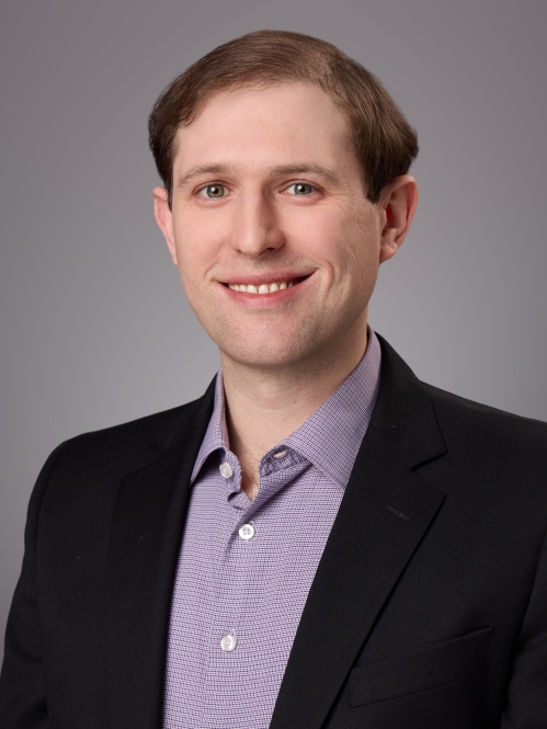 Headshot of male with short brown wavy hair wearing a black suit jacket with a patterned purple button down shirt.