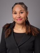 Headshot of Black woman with long dark brown hair wearing gold hoop earrings, a black shirt with a black suit jacket. 