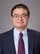 Headshot of male with short black hair and eyeglasses wearing a dark blue suit jacket, a light blue button down shirt, and a burgundy tie.