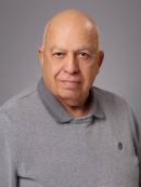 Headshot of bald male wearing a grey polo shirt underneath a grey pullover sweater.