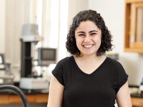 White, female college student poses in a lab. She is smiling broadly wearing a black short sleeve shirt. She has black curly hair.