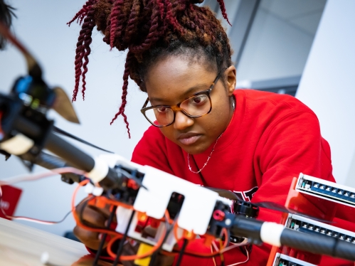 Black female engineering student works on a device. She wears a red sweatshirt, has glasses, and braids in a topknot bun.