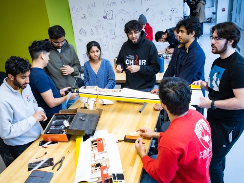 Group of nine students, eight men and one woman, work on a drone wing on a wooden lab table.