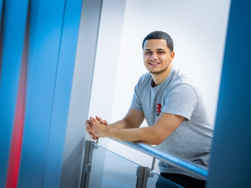 White male student leans against a railing and smiles. He has black hair and is wearing a gray Rutgers T-shirt.