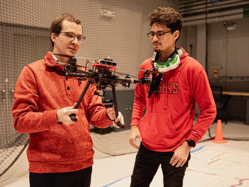 White male professer holds a drone and speaks to a white male students who is wearing a VR control headset around his neck. Both have on red shirts.