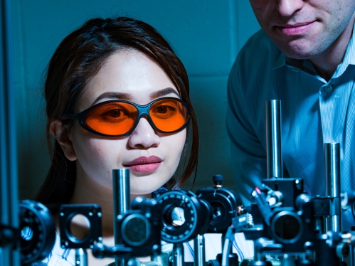 White female student wearing safety goggles with orange lenses works at laser table with a white male professor also wearing safety goggles.