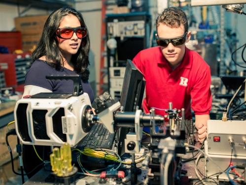 Two students wearing protective eyewear work in a laser lab. Female has black hair is on the left. Male student is wearing a read Rutgers shirt.