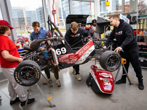Four male students work on a Formula race car in a workshop with glass walls.