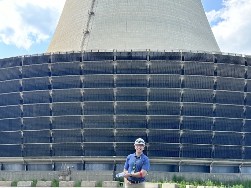 White male student poses in front of a nuclear power plant wearing a hard hat and blue polo shirt.