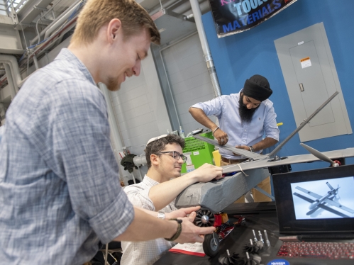 Three male students work on a airplane model in a lab. There is a computer monitor that shows a rendering of the plane 