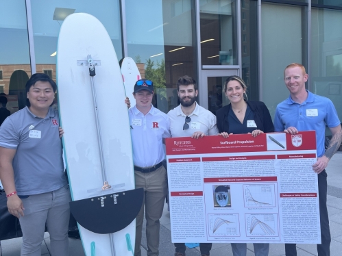 Four male students and one female student stand with their surfboard project and a senior design poster.