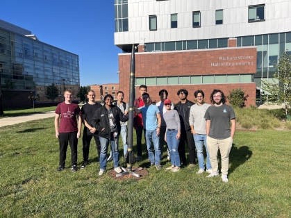 11 engineering students stand holding a rocket outside