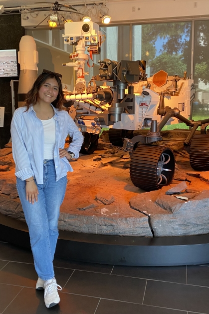 Young college student stands with one hand on her hip posing in front of a lunar rover. She is wearing jeans, a light blue blouse unbuttoned over a white shirt. She has long dark hair.