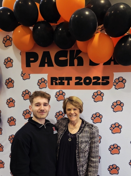Male student standing on left side and female professor standing on right side in front of a photo backdrop with orange paw prints