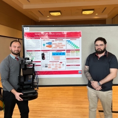 Two male students stand in front of a poster. The student on the left holds a robot while the other student stands with his arms. crossed.