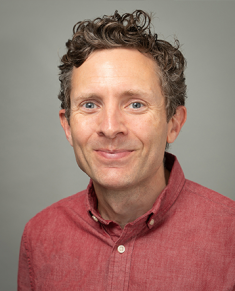 Head shot of male with short, curly hair, wearing a light red button down shirt.