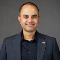 Head shot of male, Bhanu Sood, wearing a dark grey suit and a black button down shirt.
