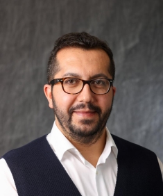 Headshot of male with short black hair, a beard and moustache, wearing eyeglasses a white button down dress shirt with a black vest.