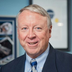 Headshot of male with short grey hair, wearing a dark suit jacket with a blue shirt and patterned tie.