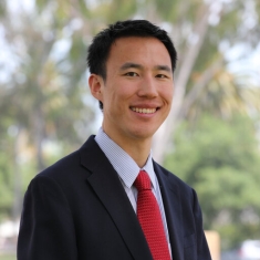 Headshot of male with black, short hair, wearing a black suit, stipped blue and white button down shirt, and a red tie.