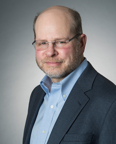 Headshot of balding male professor wearing eyeglasses, a dark suit jacket, and a button down blue shirt.